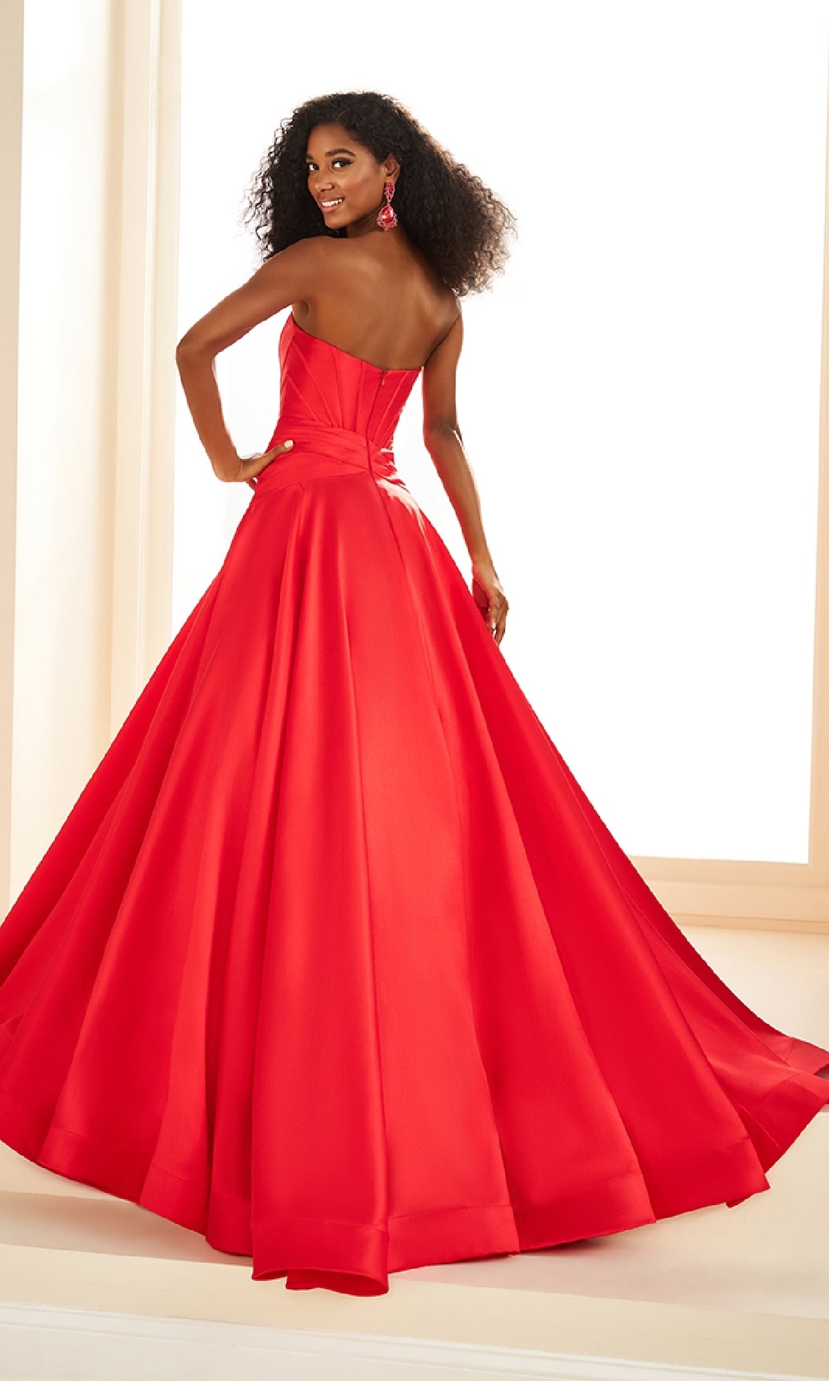 A woman in the Ellie Wilde EW36105 long prom dress—a strapless mikado ball gown with a corset bodice—poses with her back to the camera, glancing over her shoulder in a brightly lit room.